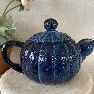 May include: A dark blue ceramic teapot with a textured surface and a shell-shaped lid handle. The teapot has a rounded body with a spout and a curved handle. The teapot is sitting on a light-colored surface.