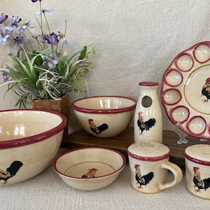 May include: A collection of cream-colored ceramic kitchenware with a rooster motif. The set includes bowls, mugs, a milk bottle, and an egg plate. Each piece has a red rim and a rooster illustration in black, red, and yellow.