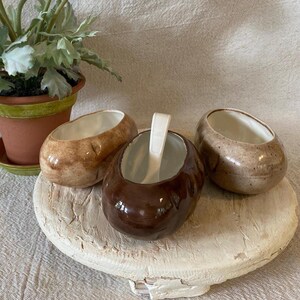 May include: Three oval-shaped ceramic bowls with a glossy finish, featuring a light tan bowl, a medium brown bowl, and a dark brown bowl. The dark brown bowl contains a white ceramic spoon. The bowls are arranged on a round, textured white base.