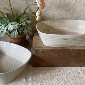 May include: Two off-white ceramic serving bowls. One is rectangular with a ribbed design and a small decorative mark. The other is square with a spiral pattern. A potted plant with pink flowers is in the background.