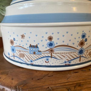 May include: Cream-colored ceramic casserole dish with a blue band and lid. The dish features a pastoral scene with houses, trees, and sheep. The design includes blue hearts and brown accents. Handles are on either side.