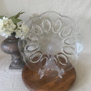 May include: Clear glass egg serving platter with 12 oval indentations. The platter is on a clear plastic stand, and sits on a wooden surface. A decorative vase with white flowers is in the background.