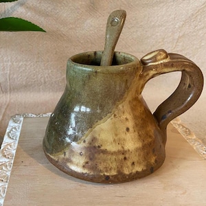 May include: A handmade ceramic mug with a matching spoon. The mug is a mix of brown and green tones, with a rounded body and a large handle. The spoon rests inside the mug, which is sitting on a wooden tray.