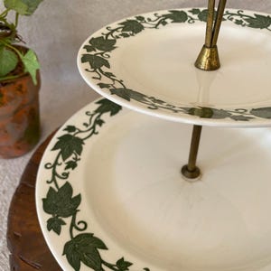 May include: Two-tiered serving tray with white plates and a green leaf design around the edges. The stand is gold-colored. A small potted plant is visible in the background.