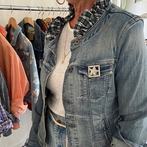 Peut inclure: Une veste en jean bleu clair avec un col &agrave; volants &agrave; carreaux verts et bleu marine, une broche en forme d'&eacute;toile argent&eacute;e sur la poche poitrine et des boutons en m&eacute;tal. La veste est port&eacute;e sur un haut blanc uni avec un collier dor&eacute; fin.