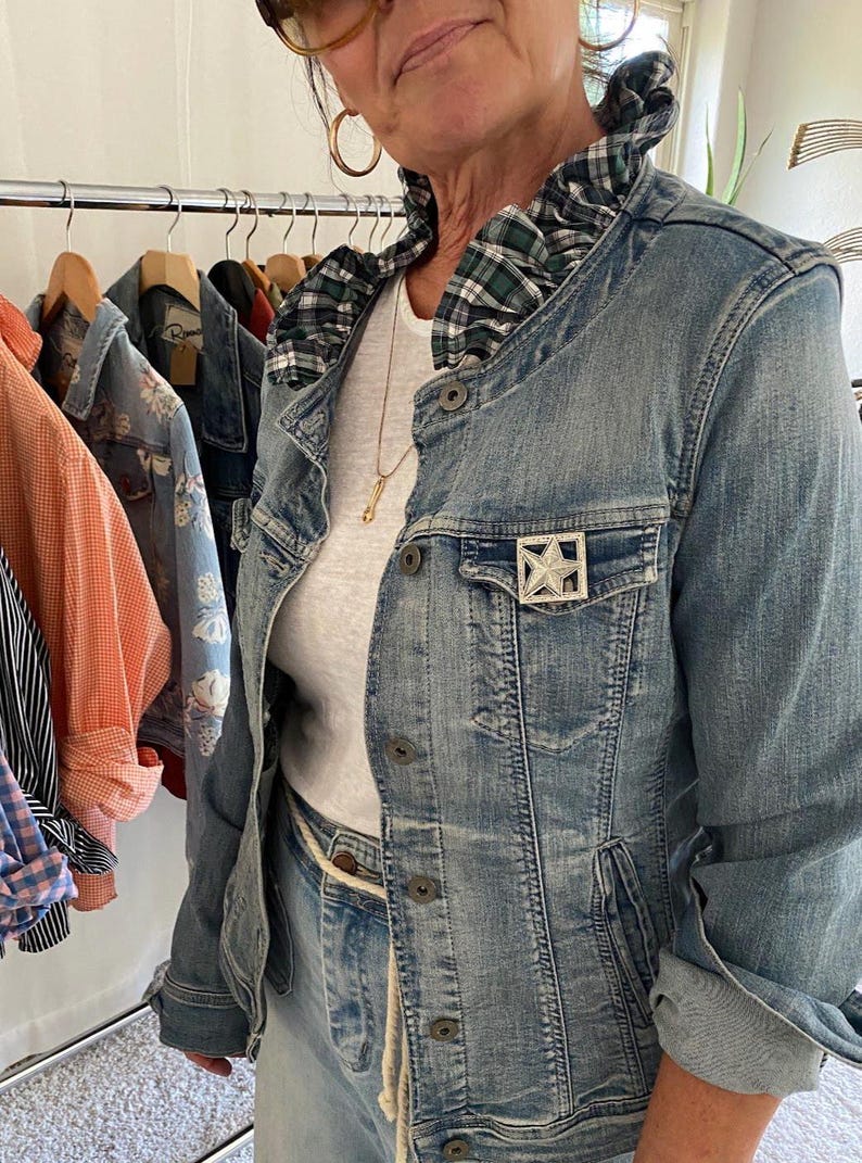 Peut inclure: Une veste en jean bleu clair avec un col &agrave; volants &agrave; carreaux verts et bleu marine, une broche en forme d'&eacute;toile argent&eacute;e sur la poche poitrine et des boutons en m&eacute;tal. La veste est port&eacute;e sur un haut blanc uni avec un collier dor&eacute; fin.