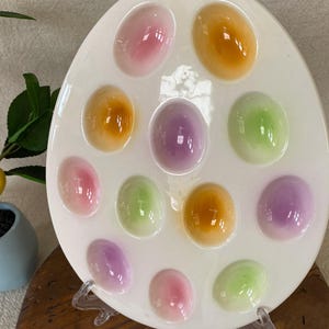 May include: White ceramic egg-shaped serving platter with 13 oval indentations, each holding a colorful, translucent egg-shaped object. Colors include pink, orange, green, and purple. The platter is propped up on a clear stand.