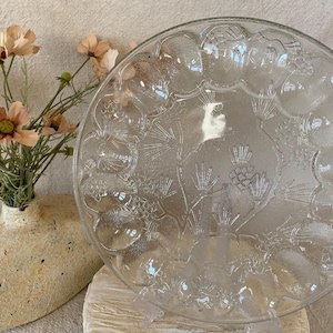 May include: A clear glass serving plate with a decorative embossed design of pinecones and foliage. The plate is round and sits on a clear plastic stand. A vase with peach-colored flowers is in the background.