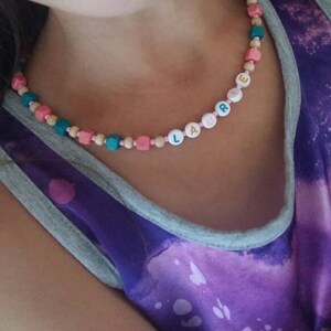 以下が含まれることがあります： 紫とグレーのタンクトップを着た女の子のクローズアップ。木製のビーズネックレスには、金色のアクセントがついた白い文字で「LAURE」という名前が書かれています。