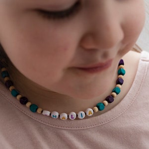 以下が含まれることがあります： ピンクのシャツを着た若い女の子のクローズアップ。カラフルな文字で「HALIA」と綴られた木製のビーズネックレスをしています。
