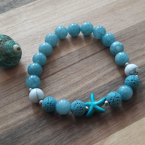 May include: A beaded bracelet featuring light blue and white round beads, interspersed with textured blue beads and a turquoise starfish charm. A teal seashell sits nearby on a wooden surface. The bracelet is approximately 7 inches in circumference.