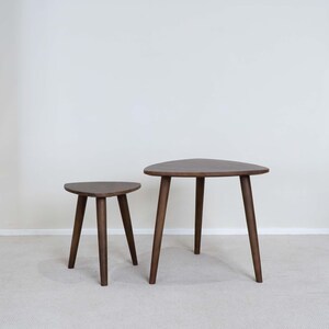 May include: A set of two dark brown wooden side tables with rounded triangular tops and three tapered legs each. The tables are of different heights, with the smaller table positioned to the left of the larger table, both featuring a smooth finish.