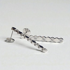 May include: Silver earrings with a linear design. Each earring has a series of hexagonal shapes, each with a small, sparkling gem. The earrings have a post and backing for secure wear. The background is white.