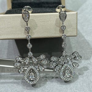 May include: A pair of elegant silver earrings with a floral design. Each earring features a teardrop-shaped stud, a series of round stones, and a cluster of smaller stones. The bottom of each earring has a large teardrop-shaped stone.