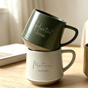Può includere: Tre tazze in ceramica con manico, caratterizzate da testo personalizzato. Una tazza verde oliva è impilata su una tazza color crema, entrambe con nomi e date. Una terza tazza beige maculata è posta accanto, anch'essa con nome e data in corsivo.