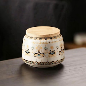 May include: A decorative ceramic jar with a wooden lid. The jar is white with a repeating pattern of blue, gold, and black floral designs. The jar sits on a dark wooden surface.