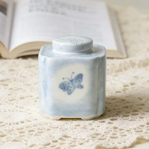 May include: A square, light blue ceramic jar with a lid, featuring a blue butterfly design. The jar is on a cream-colored lace tablecloth, with an open book in the background. The jar is approximately 4 inches tall.