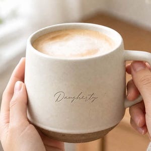 Può includere: Una tazza in ceramica color crema con una consistenza maculata e una base marrone scuro. La tazza presenta il nome Dougherty scritto in un sottile corsivo scuro sulla parte anteriore ed è riempita con una bevanda al caffè marrone chiaro con schiuma.