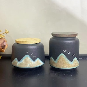 May include: Two black ceramic jars with wooden lids. The jars feature a mountain design in shades of tan and teal, with small white bird silhouettes. One jar has a bamboo lid, the other has a dark wood lid.
