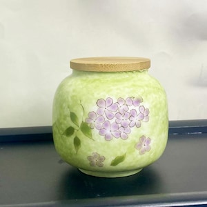 K&ouml;nnte beinhalten: Ein runder, hellgr&uuml;ner Keramiktopf mit Holzdeckel. Der Topf ist mit einer Ansammlung von lila Blumen und gr&uuml;nen Bl&auml;ttern verziert. Der Topf ist etwa 13 cm hoch.