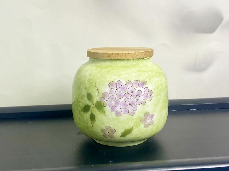 K&ouml;nnte beinhalten: Ein runder, hellgr&uuml;ner Keramiktopf mit Holzdeckel. Der Topf ist mit einer Ansammlung von lila Blumen und gr&uuml;nen Bl&auml;ttern verziert. Der Topf ist etwa 13 cm hoch.