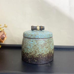 May include: A ceramic jar with a lid, featuring a speckled turquoise and brown glaze. The jar has a bamboo-style handle on the lid, and the overall design suggests a rustic or artisanal aesthetic. The jar is likely used for storage.