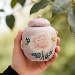 May include: A small, round, light purple ceramic jar with a lid, held in a hand. The jar is decorated with a large, pink and white peony flower design, with green and blue leaves. The jar is likely for decorative purposes.