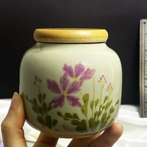 K&ouml;nnte beinhalten: Ein kleines, rundes Keramikgef&auml;&szlig; mit Holzdeckel. Das Gef&auml;&szlig; ist mit lila Blumen und gr&uuml;nem Laub bemalt. Der Text auf dem Schild im Hintergrund lautet: "I SEE TREES OF GREEN RED ROSES TOO I SEE THEM BLOOM FOR ME AND YOU."