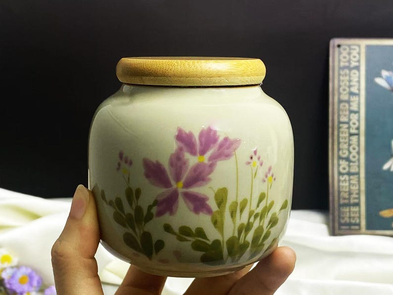 K&ouml;nnte beinhalten: Ein kleines, rundes Keramikgef&auml;&szlig; mit Holzdeckel. Das Gef&auml;&szlig; ist mit lila Blumen und gr&uuml;nem Laub bemalt. Der Text auf dem Schild im Hintergrund lautet: "I SEE TREES OF GREEN RED ROSES TOO I SEE THEM BLOOM FOR ME AND YOU."