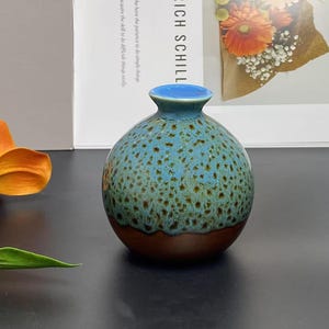 May include: A small, round ceramic vase with a speckled turquoise and brown glaze. The vase has a narrow neck and a dark brown base. The background includes a flower and a framed print.