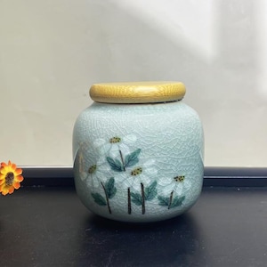 K&ouml;nnte beinhalten: Hellblaue Keramikdose mit Holzdeckel. Die Dose hat ein Blumendesign mit wei&szlig;en Bl&uuml;ten und gr&uuml;nen Bl&auml;ttern. Die Dose hat eine Craquel&eacute;-Glasur, und der Deckel ist aus hellem Holz gefertigt.