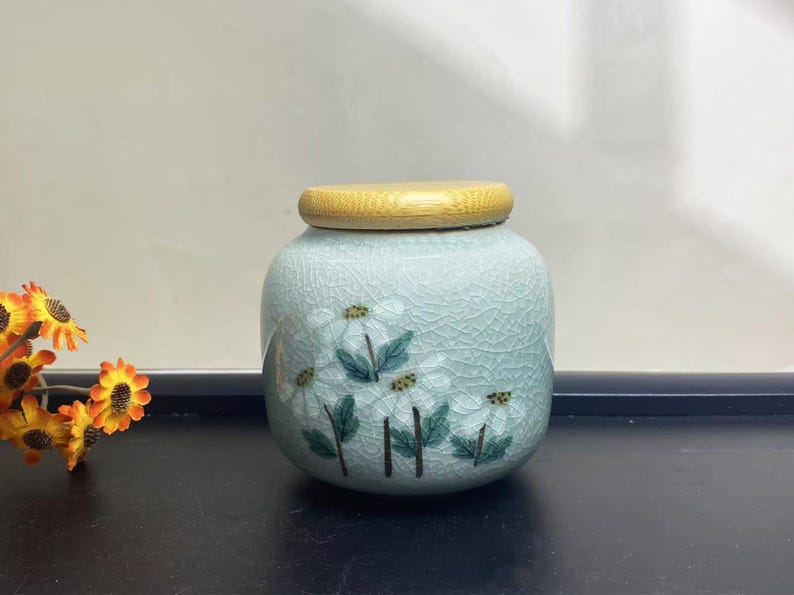 K&ouml;nnte beinhalten: Hellblaue Keramikdose mit Holzdeckel. Die Dose hat ein Blumendesign mit wei&szlig;en Bl&uuml;ten und gr&uuml;nen Bl&auml;ttern. Die Dose hat eine Craquel&eacute;-Glasur, und der Deckel ist aus hellem Holz gefertigt.