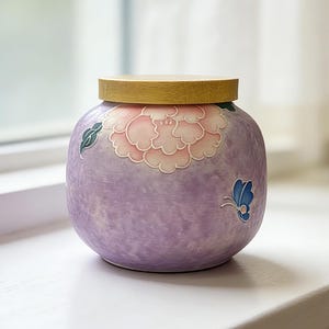 May include: A round, lavender ceramic jar with a wooden lid. The jar is decorated with a pink and white floral design and a blue butterfly. The jar is sitting on a white surface.