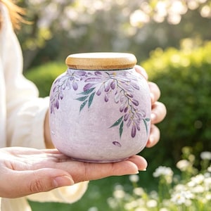 May include: A round ceramic jar with a light purple textured finish, featuring hand-painted purple wisteria flowers and green leaves. The container is topped with a smooth, circular light brown wooden lid that sits flush on the top opening.