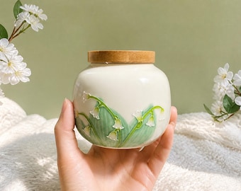 May include: A round, cream-colored ceramic jar featuring a raised, embossed design of green lily of the valley leaves and white bell-shaped flowers. The container is topped with a circular, light-brown wooden lid that fits flush with the rim.
