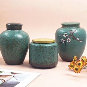 May include: Three teal ceramic jars with lids. The jars have a crackle glaze finish. One jar has a black lid, one has a gold lid, and the third has a floral design. A bouquet of yellow and orange flowers is in the foreground.
