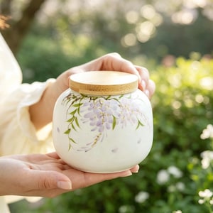 Può includere: Un barattolo in ceramica bianca di forma rotonda con un disegno dipinto a mano di fiori di glicine viola chiaro e foglie verdi. Il contenitore è sormontato da un coperchio piatto e circolare in legno marrone chiaro.