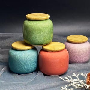 May include: Four ceramic jars in green, blue, coral, and pink, each with a wooden lid. The jars have a crackle glaze finish. A rose and decorative sprig are in the bottom right corner.