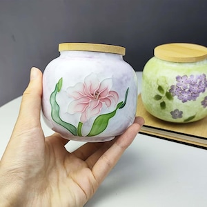 May include: A hand-painted ceramic jar with a light pink and white floral design, featuring a pink flower and green leaves. The jar has a wooden lid. Another jar with a green background and purple flowers is also visible.