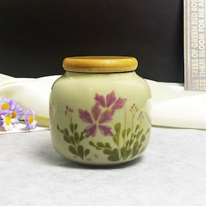 K&ouml;nnte beinhalten: Ein kleines, rundes Keramikgef&auml;&szlig; in Cremefarbe mit einem Holzdeckel. Das Gef&auml;&szlig; ist mit lila floralen Designs und gr&uuml;nen Bl&auml;ttern verziert. Das Gef&auml;&szlig; steht auf einer wei&szlig;en Oberfl&auml;che.