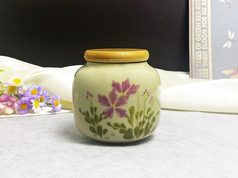 K&ouml;nnte beinhalten: Ein kleines, rundes Keramikgef&auml;&szlig; in Cremefarbe mit einem Holzdeckel. Das Gef&auml;&szlig; ist mit lila floralen Designs und gr&uuml;nen Bl&auml;ttern verziert. Das Gef&auml;&szlig; steht auf einer wei&szlig;en Oberfl&auml;che.