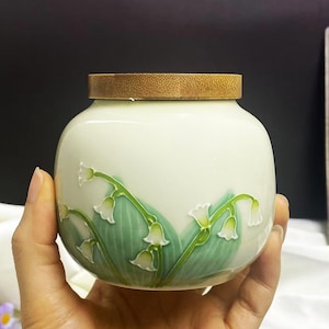 May include: A small, off-white ceramic jar with a wooden lid. The jar is decorated with a green and white lily of the valley design. The jar is held in a hand.