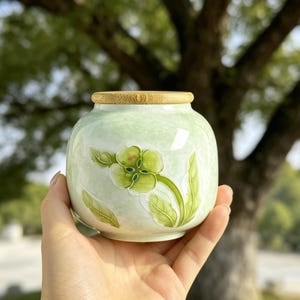 Peut inclure: Petit pot rond en céramique vert clair avec un couvercle en bambou. Le pot présente un motif floral dans les tons de vert et de blanc. Le pot est tenu dans une main sur un fond flou d'arbres.