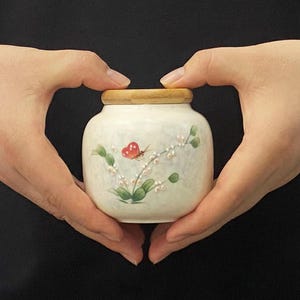 K&ouml;nnte beinhalten: Ein kleines, cremefarbenes Keramikgef&auml;&szlig; mit Holzdeckel. Das Gef&auml;&szlig; ist mit einem gemalten Schmetterling und einem floralen Design in Rot, Gr&uuml;n und Wei&szlig; verziert. Das Gef&auml;&szlig; wird von zwei H&auml;nden gehalten.