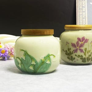 K&ouml;nnte beinhalten: Zwei Keramikgl&auml;ser mit Holzdeckeln. Ein Glas zeigt ein Maigl&ouml;ckchen-Design in Gr&uuml;n und Wei&szlig;. Das andere Glas hat ein Blumendesign in Lila und Gr&uuml;n. Beide Gl&auml;ser haben einen hellgr&uuml;nen Hintergrund.