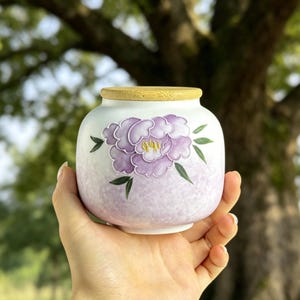 May include: A small, white ceramic jar with a bamboo lid. The jar is decorated with a large, purple peony flower design and green leaves. The jar is held in a hand against a blurred background of a tree.