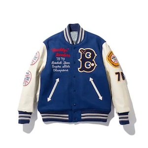 Op de afbeelding: Een blauwe varsity-jas met crèmekleurige mouwen, voorzien van een grote letter B op de borst, geborduurde tekst Brooklyn Breakers 78 79 Baseball Team Empire State Champions en diverse ronde patches op de mouwen.