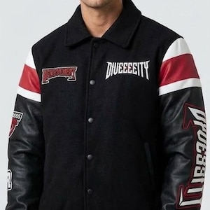 Pode incluir: Uma jaqueta varsity preta com corpo de lã e mangas de couro sintético preto. A jaqueta apresenta listras vermelhas e brancas nas mangas, o texto branco "DIVEEEEITY" no peito e emblemas bordados em vermelho com texto branco.