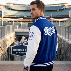 Può includere: Una giacca varsity blu e bianca con il logo Dodgers in corsivo bianco sul retro e una toppa circolare da baseball dei Dodgers sulla manica destra. La giacca presenta maniche bianche, corpo blu e polsini e colletto a coste a righe.