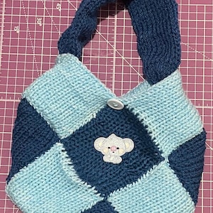 Peut inclure: Un sac fourre-tout en tricot bleu et bleu clair avec un motif à carreaux. Le sac a une seule bandoulière et une fermeture à bouton. Un petit koala brodé se trouve sur le devant du sac.